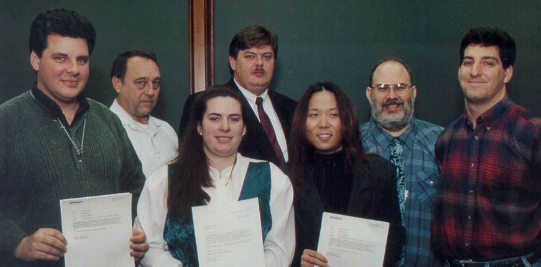 Lynn Bassford, a woman with long brown hair, poses in a vintage photo with a group of other people, several holding printed pieces of paper. Lynn wears a green velvet jumper and white blouse, and her colleagues of various ages and genders wear work clothes like suits or flannel shirts with ties. They pose in front of a dark green background.