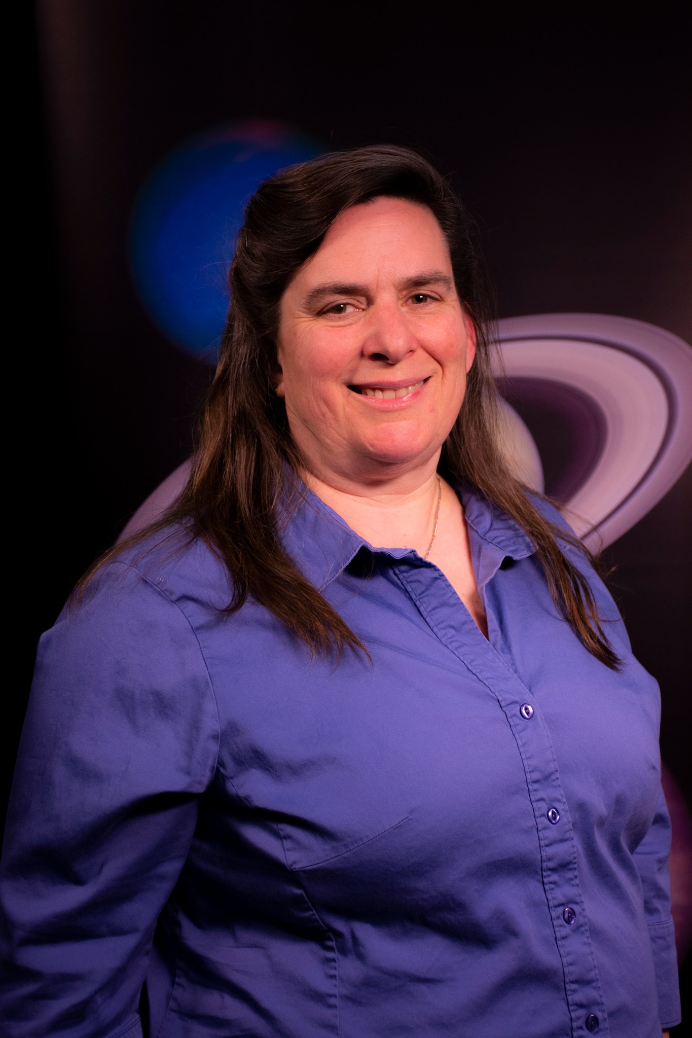 Lynn Bassford, a woman with long brown hair, smiles at the camera in an official headshot. She wears a purple collared shirt and poses in front of a photo of Saturn and Neptune.