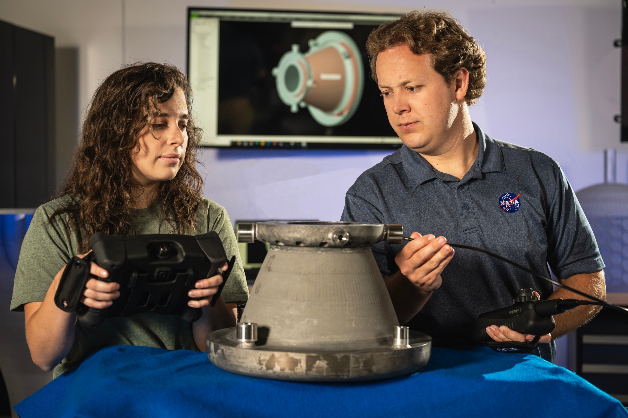 A female engineer with brown curly hair and a male engineer with short brown hair look at a nozzle on a table that has been through hot fire testing.