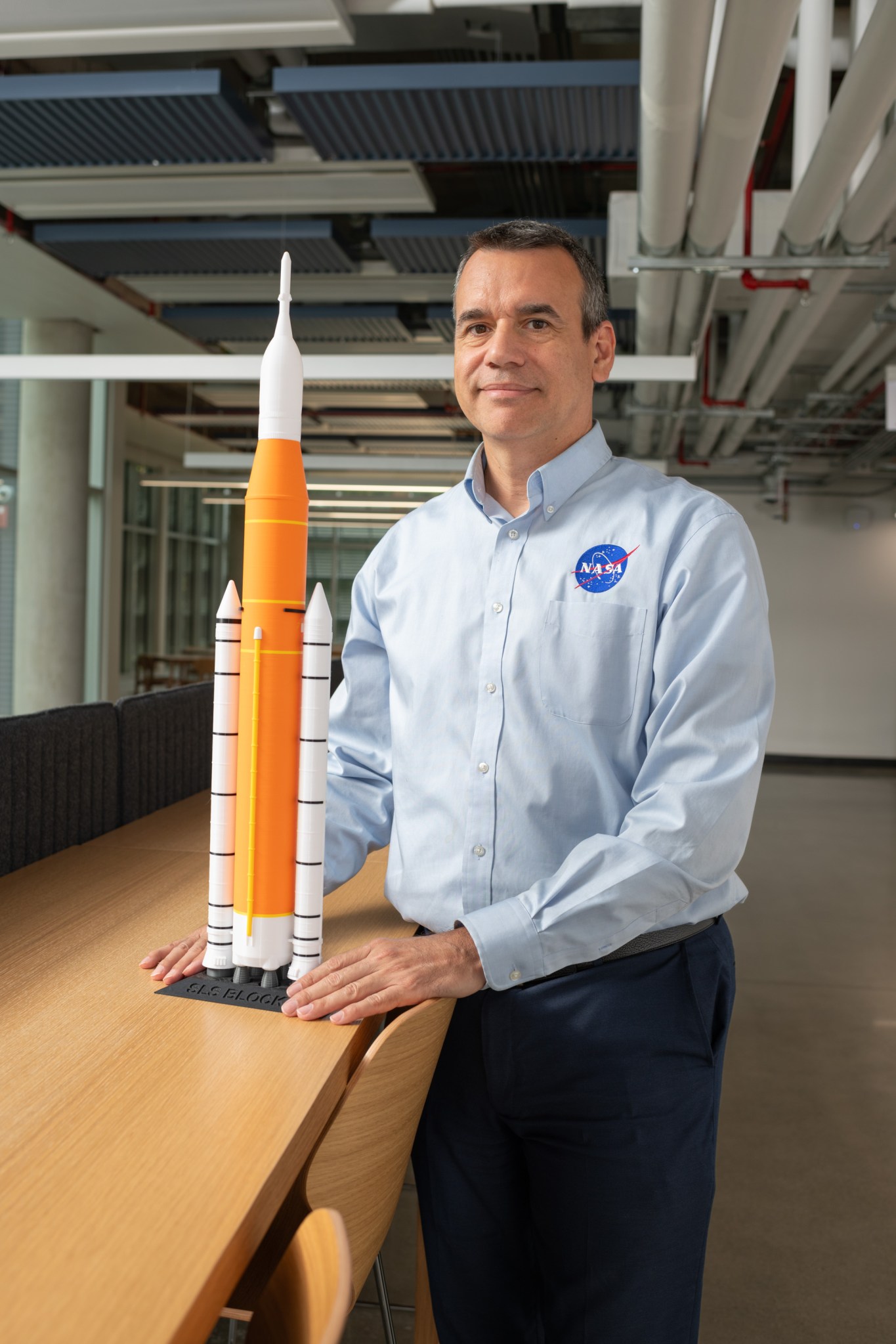Nelson Morales poses next to a small model of NASA’s Space Launch System. He is wearing a blue collared shirt with a NASA logo.