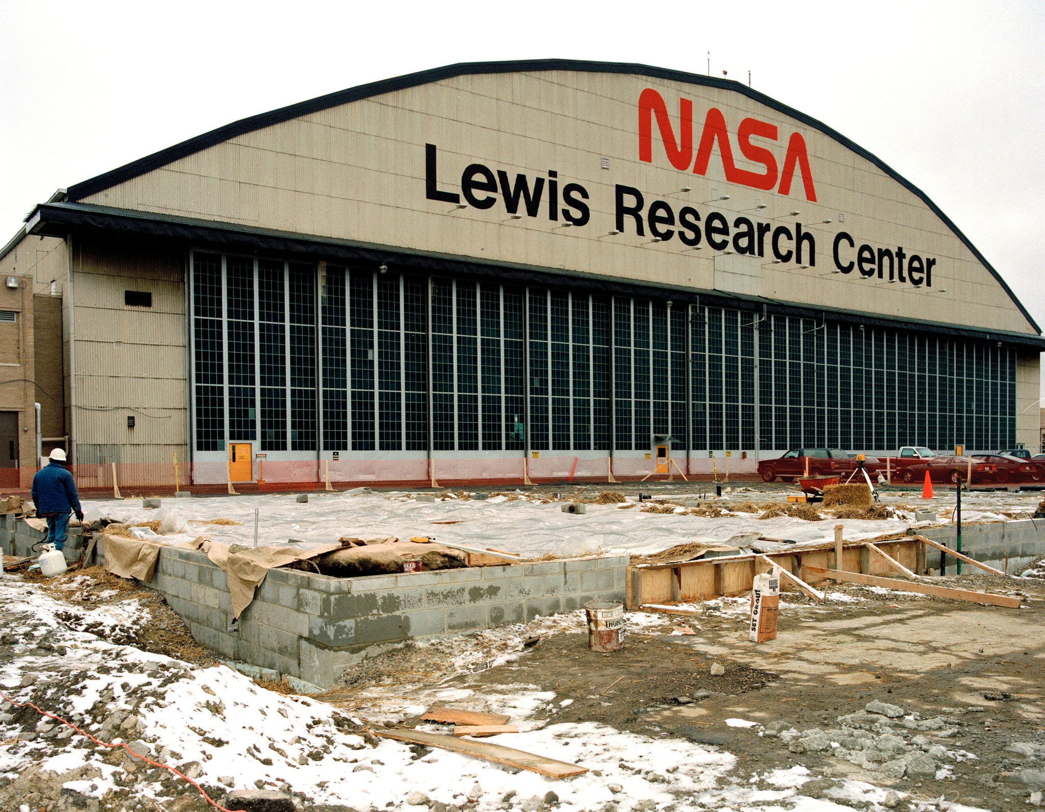 The front of the hangar at NASA's Glenn Research Center. A sign above the hangar includes a small NASA meatball insignia and large text that says, "Glenn Research Center, Lewis Field." Trees and a road are in the foreground.