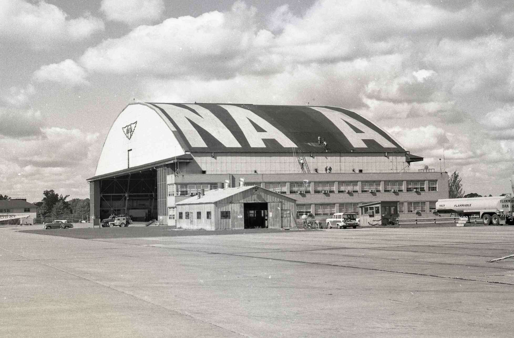 A black-and-white photo of a hangar on a cloudy day. On the hangar’s roof are huge painted letters: “N-A-A” with a space between the two A’s where a letter has been removed.