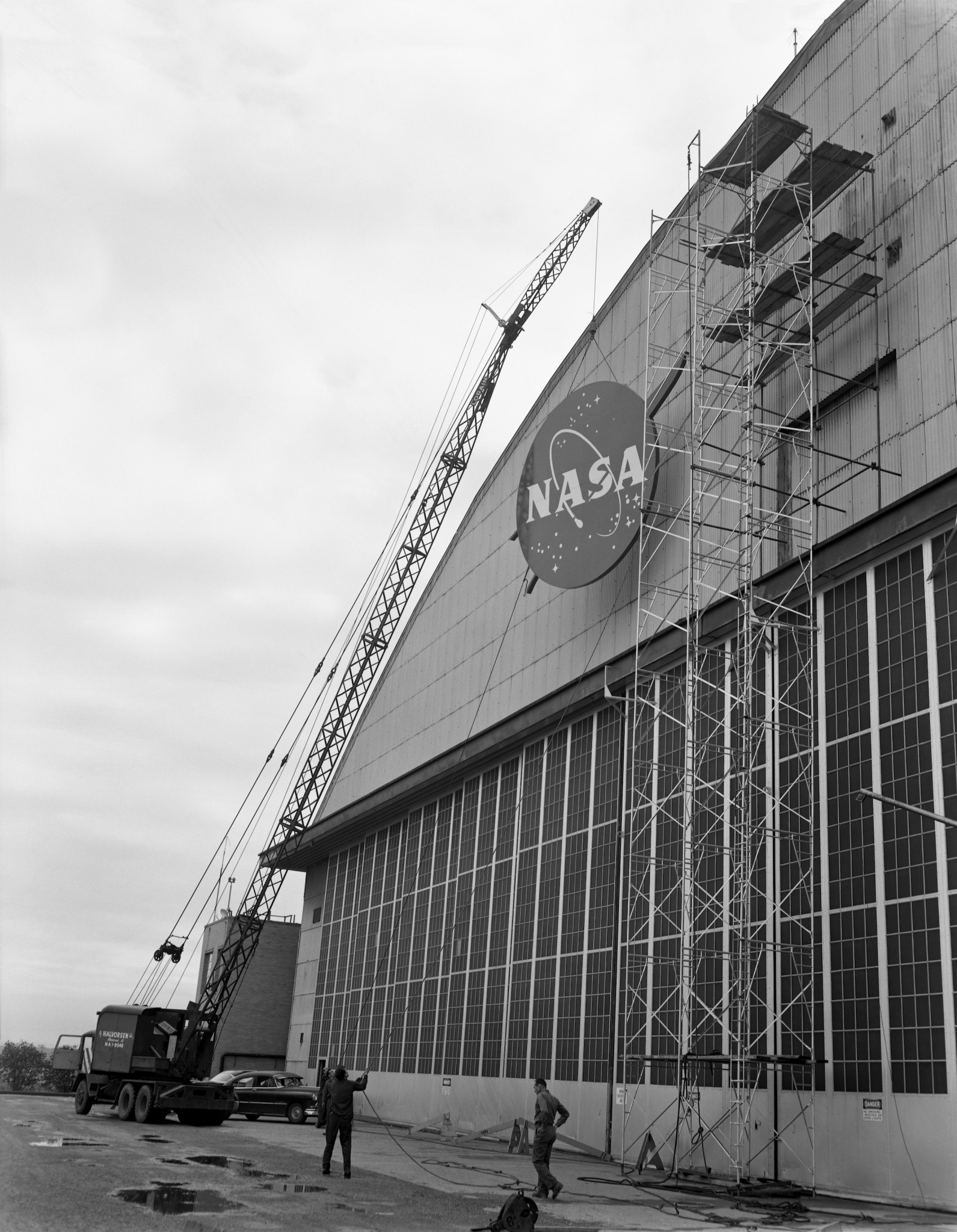 Uma foto em preto e branco de um guindaste levantando uma grande placa com a insígnia “almôndega” da NASA na parte externa de um hangar.