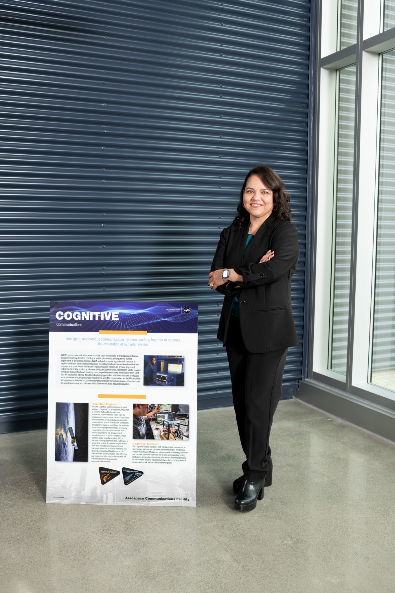 Janette C. Briones stands to the right of a poster about cognitive communications. She is crossing her arms and wearing professional attire. She poses in front of a blue metal wall.