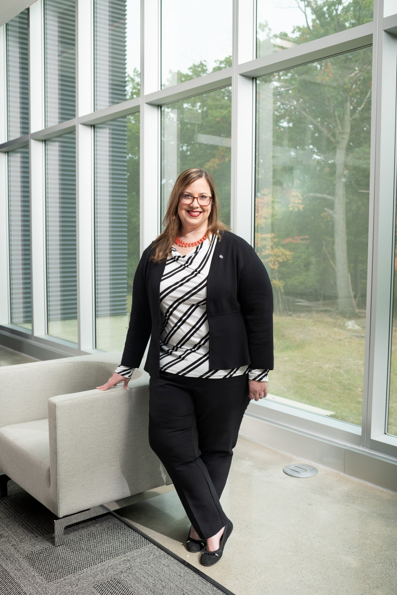 Azlin Biaggi-Labiosa stands with her hand resting on a white couch in front of a large glass window. Trees can be seen through the window, and she is wearing glasses and professional attire.