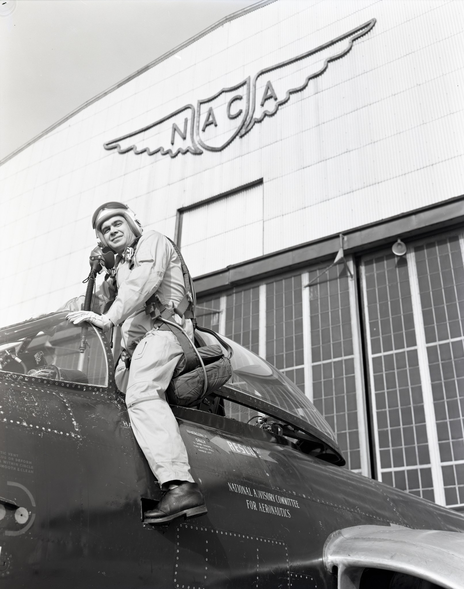 A black-and-white photo of a pilot posing as he climbs into a plane’s cockpit. Behind him in the background is a hangar with a large sign hung on it that has the letters “N-A-C-A” and a pair of wings.
