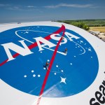 Workers paint the blue NASA “meatball” insignia on the top of NASA Glenn Research Center’s hangar roof. The workers are tethered to the roof and are very small in comparison to the large logo.