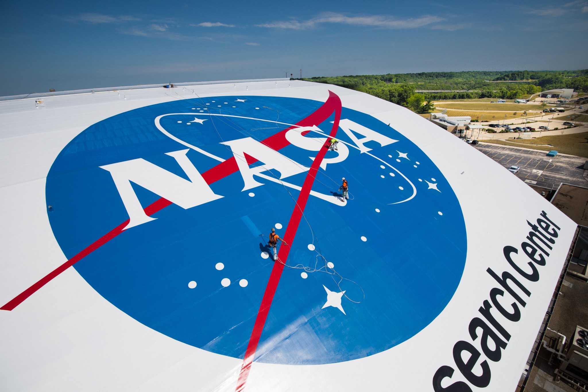 A black-and-white photo of a crane lifting a large sign with the NASA “meatball” insignia onto the outside of a hangar building.