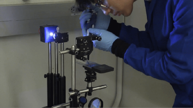 A worker wearing blue coveralls and gloves uses a syringe to place liquid on a glass surface held by a clamp. There is a black camera above the surface on another clamp in the foreground and a bright light and a focusing lens opposite the camera.