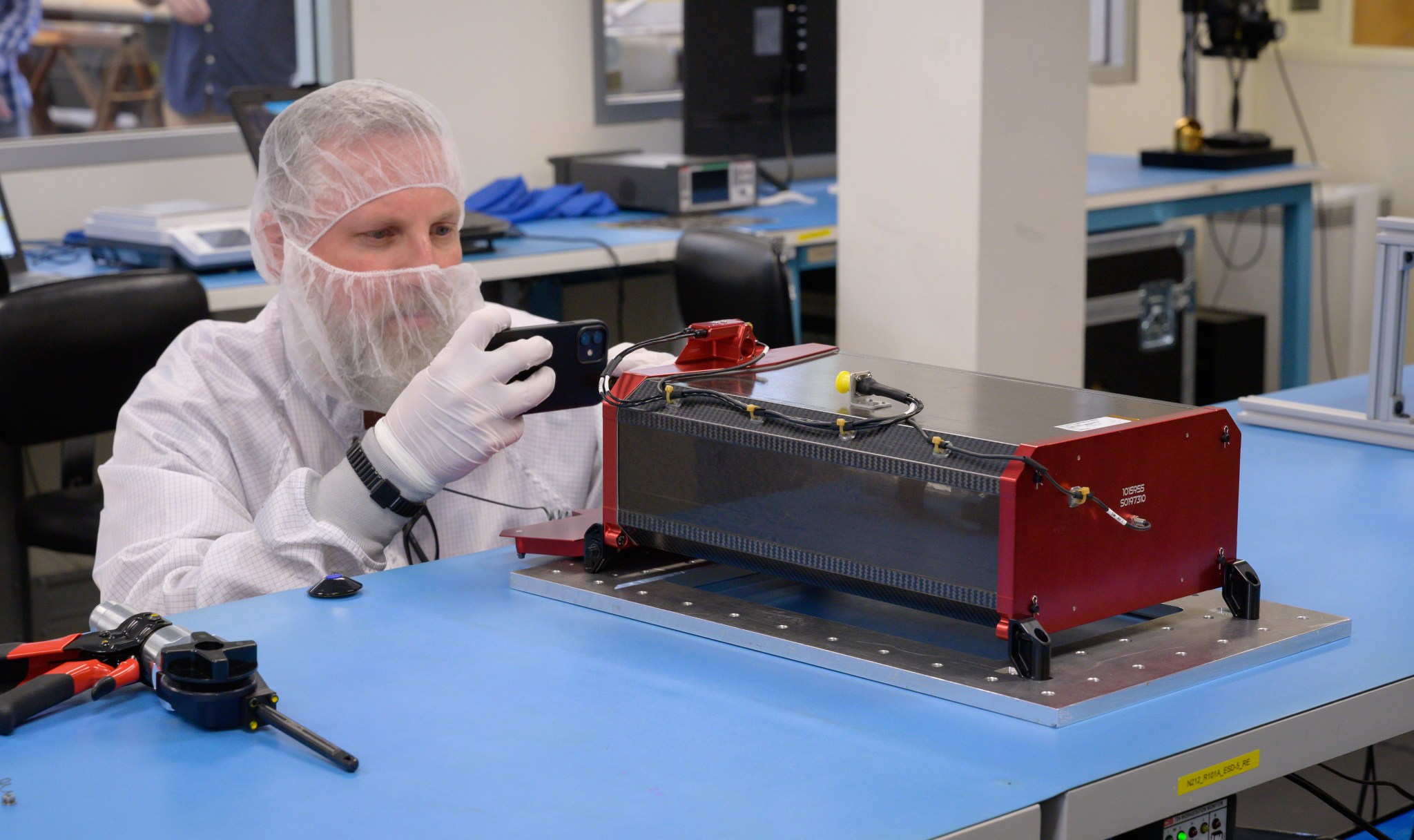 Researcher with hair net, mask, gloves, and lab coat works on box-like mechanical structure on a table.