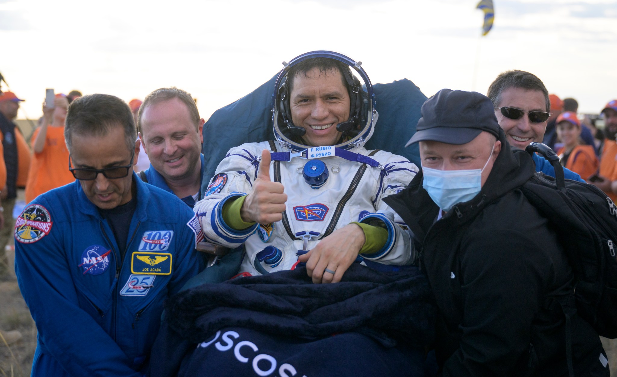 Astronaut Frank Rubio gives the camera a thumbs up as he is carried by four men, including NASA astronaut Joe Acaba (front left). Rubio wears a white spacesuit with blue accents and several mission patches. He rests against the propped-up top portion of a stretcher and has a dark blue blanket on his lap.