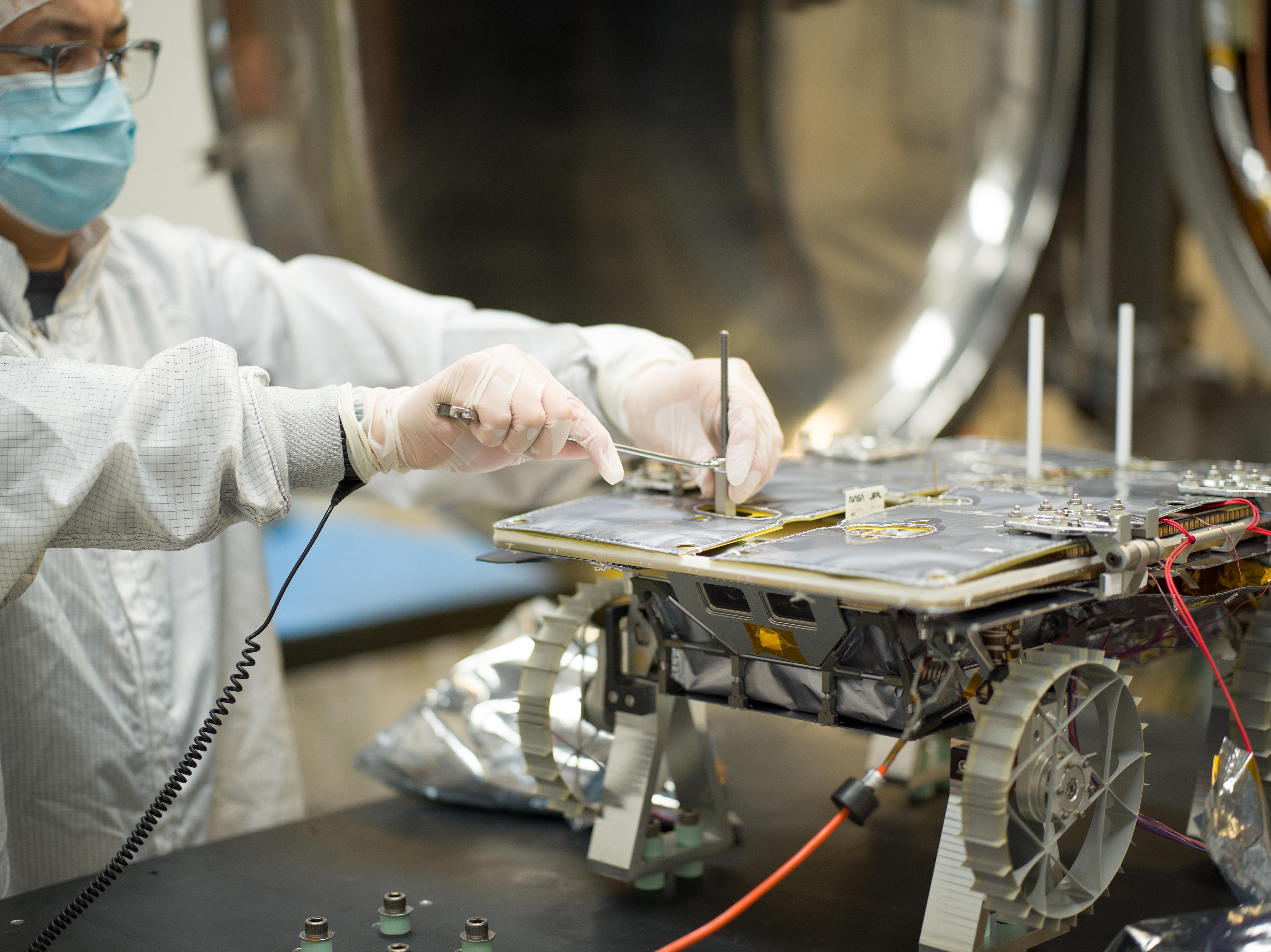 An engineer in white protective gear, a blue mask, and glasses works on a small rover. The rover is small enough to fit on a black tabletop. The rover has a flat top and four gear-like wheels.