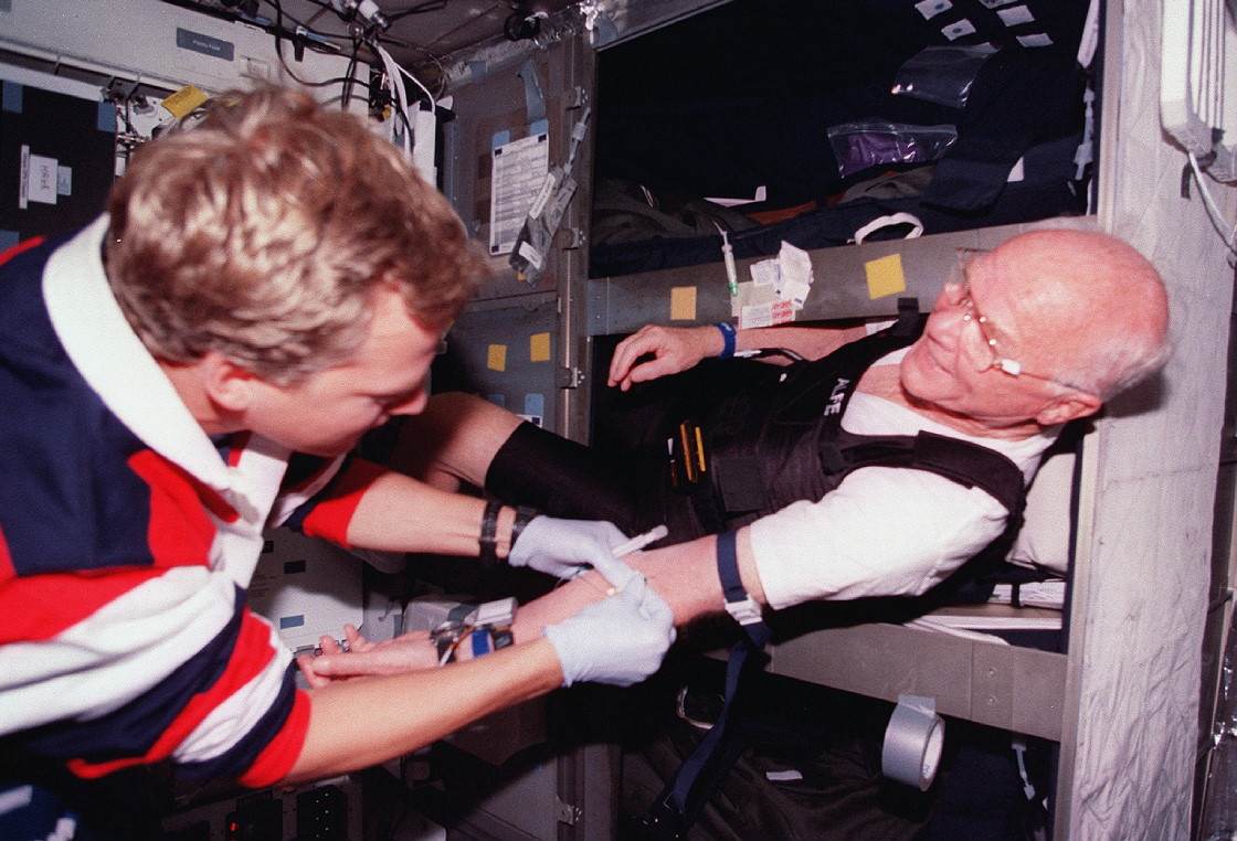 Physician astronaut Dr. Scott E. Parazynski, left, prepares to draw a blood sample from John H. Glenn