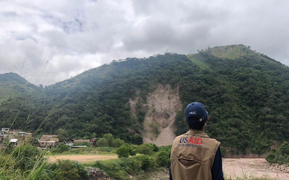 A person stands with their back to the camera, wearing a brown vest that says "USAID". They are looking at a tree-covered mountain in the distance, which has a large landslide going down it, covered in rocks, dirt, and debris. A village sits at the bottom of the hill. The sky is gray and cloudy.