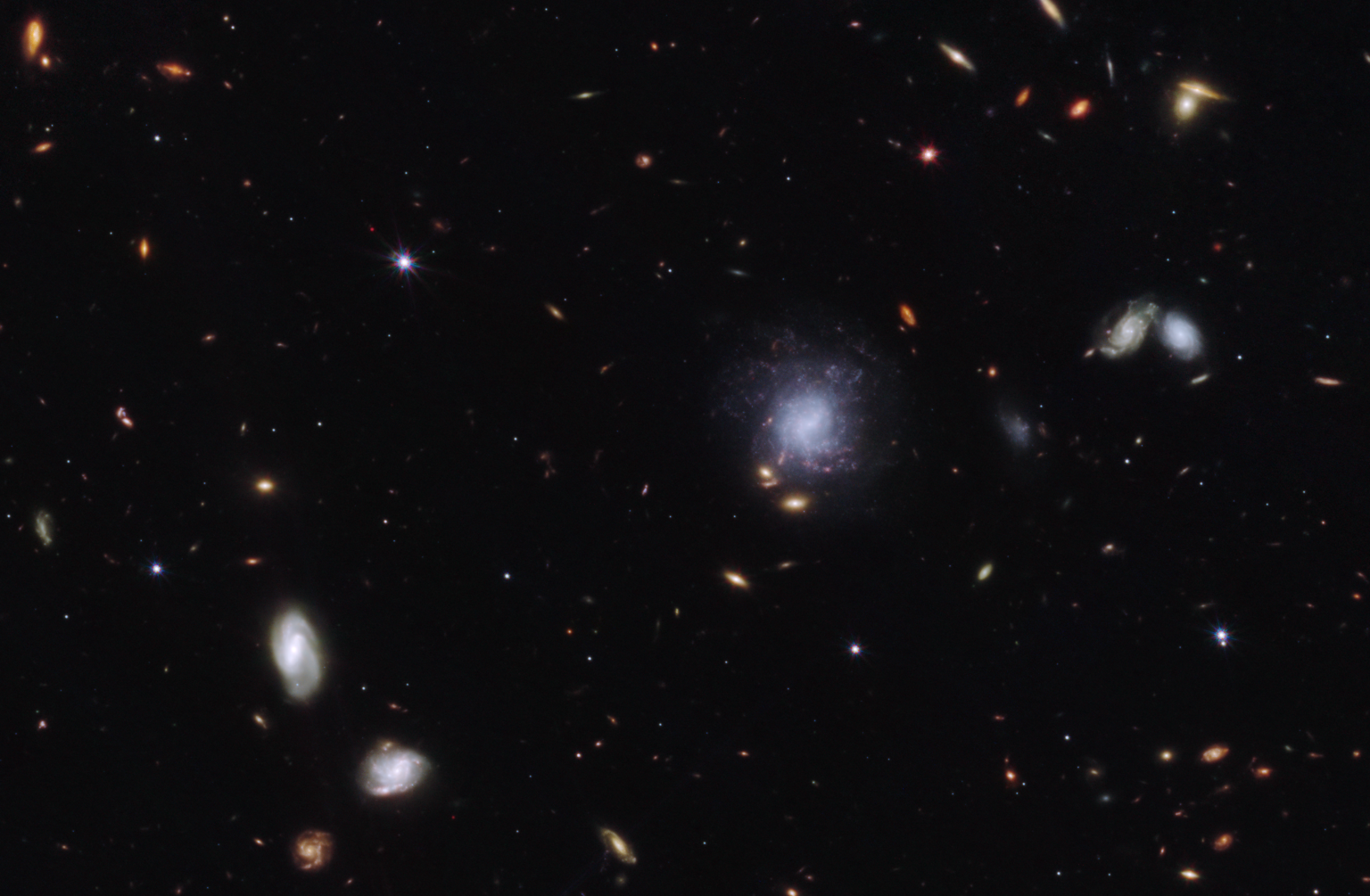 Bright galaxies and other light sources in various sizes and shapes are scattered across a black swath of space: small points, hazy elliptical-like smudges with halos, and spiral-shaped blobs. The objects vary in color: white, blue-white, yellow-white, and orange-red. Toward the center right is a blue-white spiral galaxy seen face-on that is larger than the other light sources in the image. The galaxy is labeled “former home galaxy.” Toward the upper left is a small red point, which has a white circle around it and is labeled “GRB 230307A kilonova.”