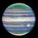 Jupiter dominates the black background of space. The image is a composite, and shows Jupiter in enhanced color, featuring the planet’s famous Great Red Spot, which appears white with light pink around the edges. The planet is striated with swirling horizontal stripes of green, periwinkle, light pink, and cream. Horizontally across the equator is a wide cream-colored band, whose height extends about 1/7 of the planet. This is the planet’s equatorial zone. The stripes across the planet interact and mix at their edges. Along both of the northern and southern poles, the planet glows in green. Bright red auroras glow just above the planet’s surface at both poles.