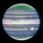 Jupiter dominates the black background of space. The image is a composite, and shows Jupiter in enhanced color, featuring the planet’s famous Great Red Spot, which appears white with light pink around the edges. The planet is striated with swirling horizontal stripes of green, periwinkle, light pink, and cream. Horizontally across the equator is a wide cream-colored band, whose height extends about 1/7 of the planet. This is the planet’s equatorial zone. The stripes across the planet interact and mix at their edges. Along both of the northern and southern poles, the planet glows in green. Bright red auroras glow just above the planet’s surface at both poles.