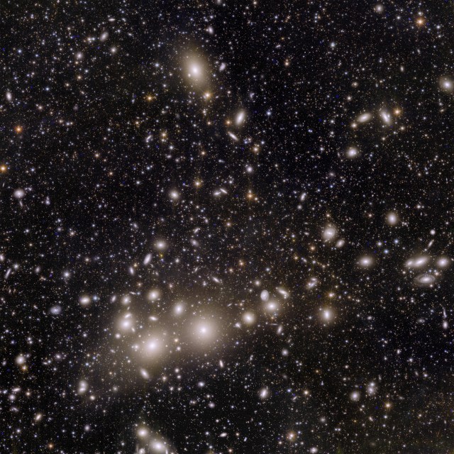 The image shows thousands of galaxies across the black expanse of space. The closest thousand or so galaxies appear as small disks of spiraling material, surrounded by halos of yellow and white light. The background is scattered with a hundred thousand more distant galaxies of different shapes, ranging in color from white to yellow to red. Most galaxies are so far away they appear as single points of light.