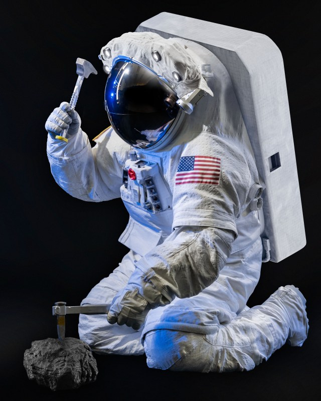 NASA engineer Scott Wray wearing NASA's xEMU (Exploration Extravehicular Mobility Unit) planetary spacesuit prototype while using a hammer and chisel to collect a rock sample.