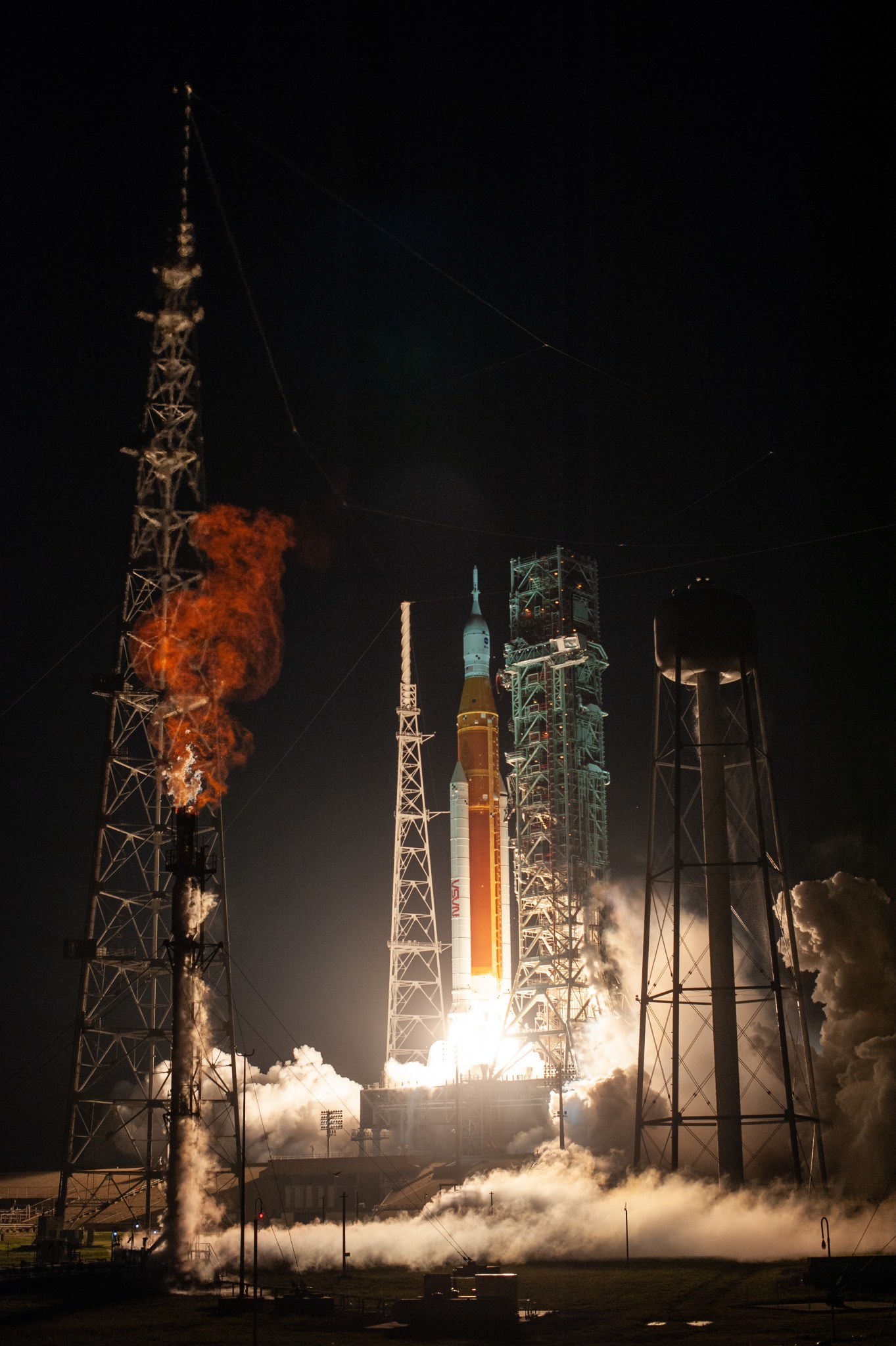 NASA's Space Launch System lifts off from the launch pad, illuminating the ground with a bright white light that bounces off of clouds of smoke. Pictured on the left side of this image is the liquid hydrogen "flare stack" where vented liquid hydrogen is burned off safely.