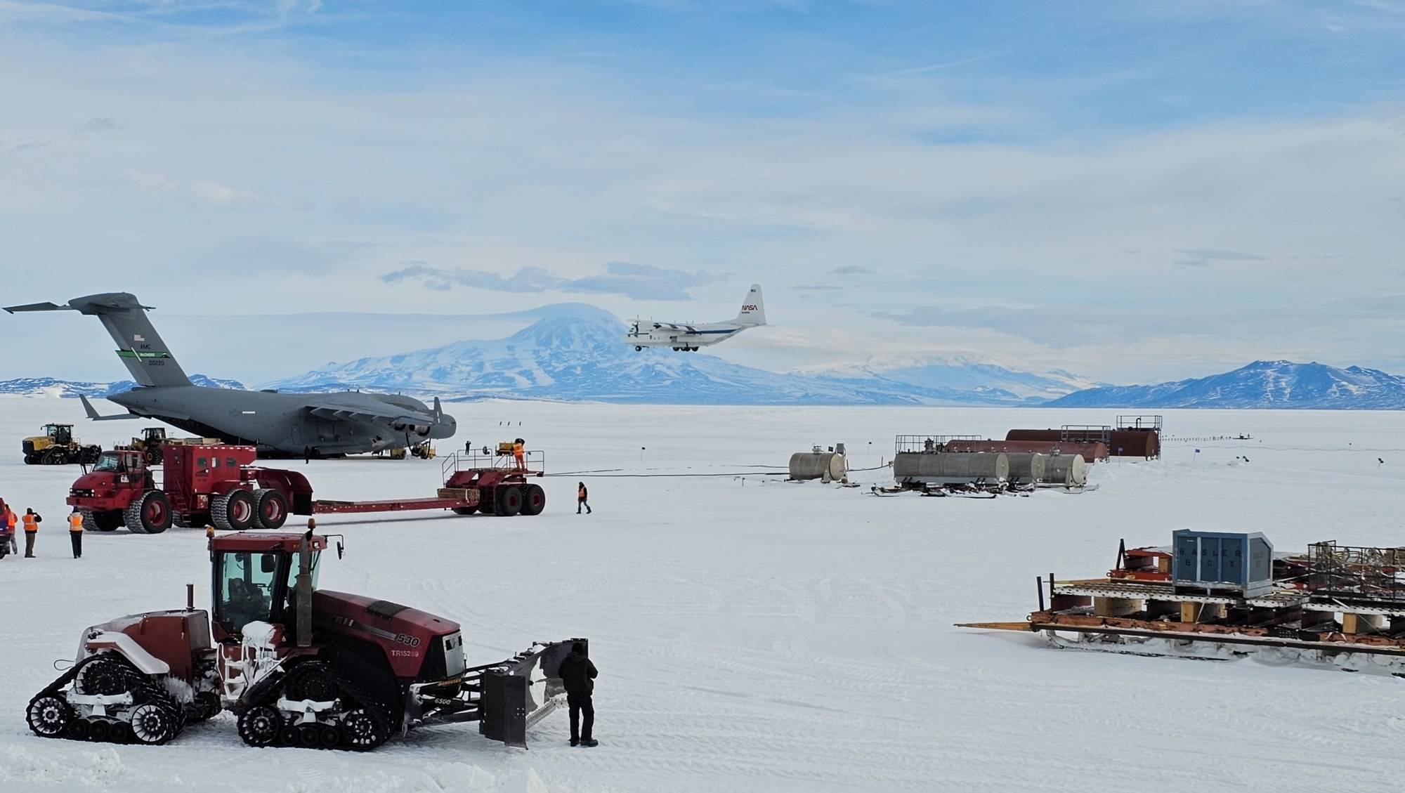 Tractors and other heavy machinery rest on the snowy plain in the foreground. In the middle of the image, a dark gray aircraft sits on the ground as a smaller white aircraft, NASA's C-130, lands.