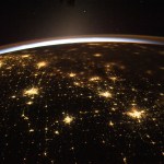 In the distance, small points of light can be seen against the darkness of space. The sun's rays peek over Earth, creating a blue haze tinged with orange. On Earth, the city lights of Chicago, Illinois, (far left) and the Dallas/Fort Worth metropolitan area (far right) shine.