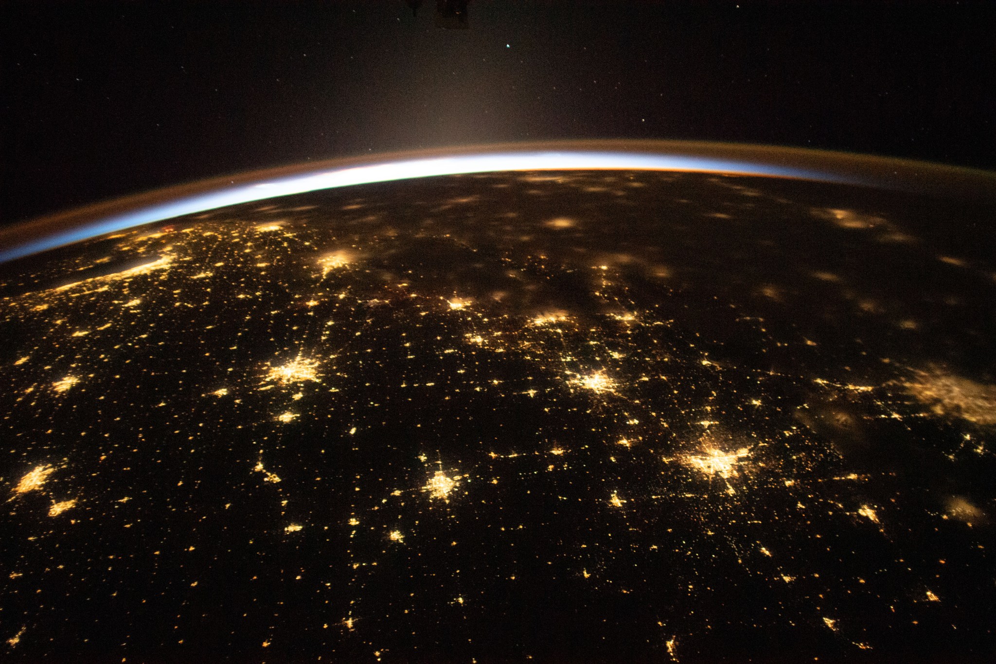 In the distance, small points of light can be seen against the darkness of space. The sun's rays peek over Earth, creating a blue haze tinged with orange. On Earth, the city lights of Chicago, Illinois, (far left) and the Dallas/Fort Worth metropolitan area (far right) shine.