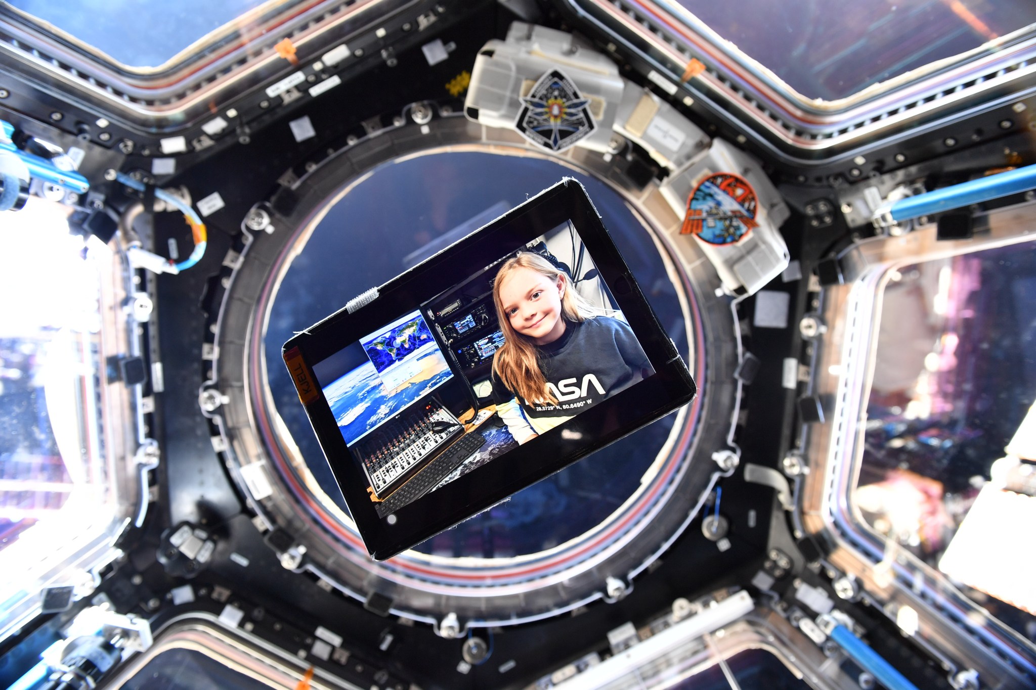 A tablet floats in the space station cupola, the screen displaying Isabella Payne in a NASA t-shirt next to a ham radio control panel and a computer showing the ISS track over the Earth. The cupola windows are behind the tablet.