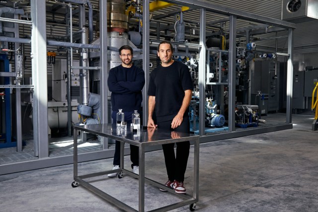 Two men stand in a room with industrial-style equipment behind them. On the metal table in front of them are four clear bottles of varying sizes with clear liquid inside the bottles. The men are wearing all black clothing and white tennis shoes.
