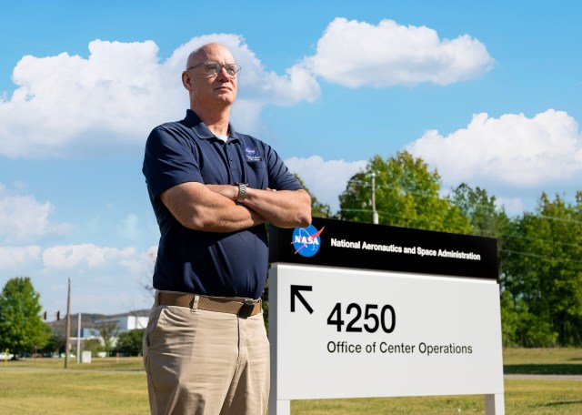 A white man with no hair stands in front of a NASA sign outdoors with his arms crossed.
