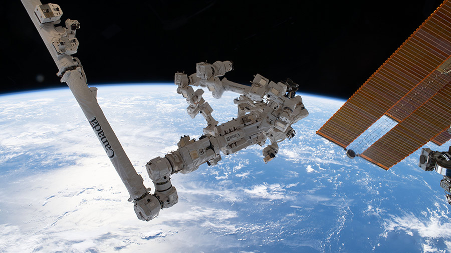 The sun's glint beams off a clear blue north Atlantic Ocean. In the foreground, are a pair of the station's main solar arrays (right) and the Canadarm2 robotic arm with its fine-tuned robotic hand Dextre (left).