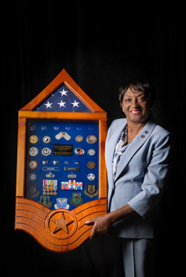 Carolyn Magsby stands next to a shadow box full of Medals. She is a Black woman wearing a great suit with a black background.
