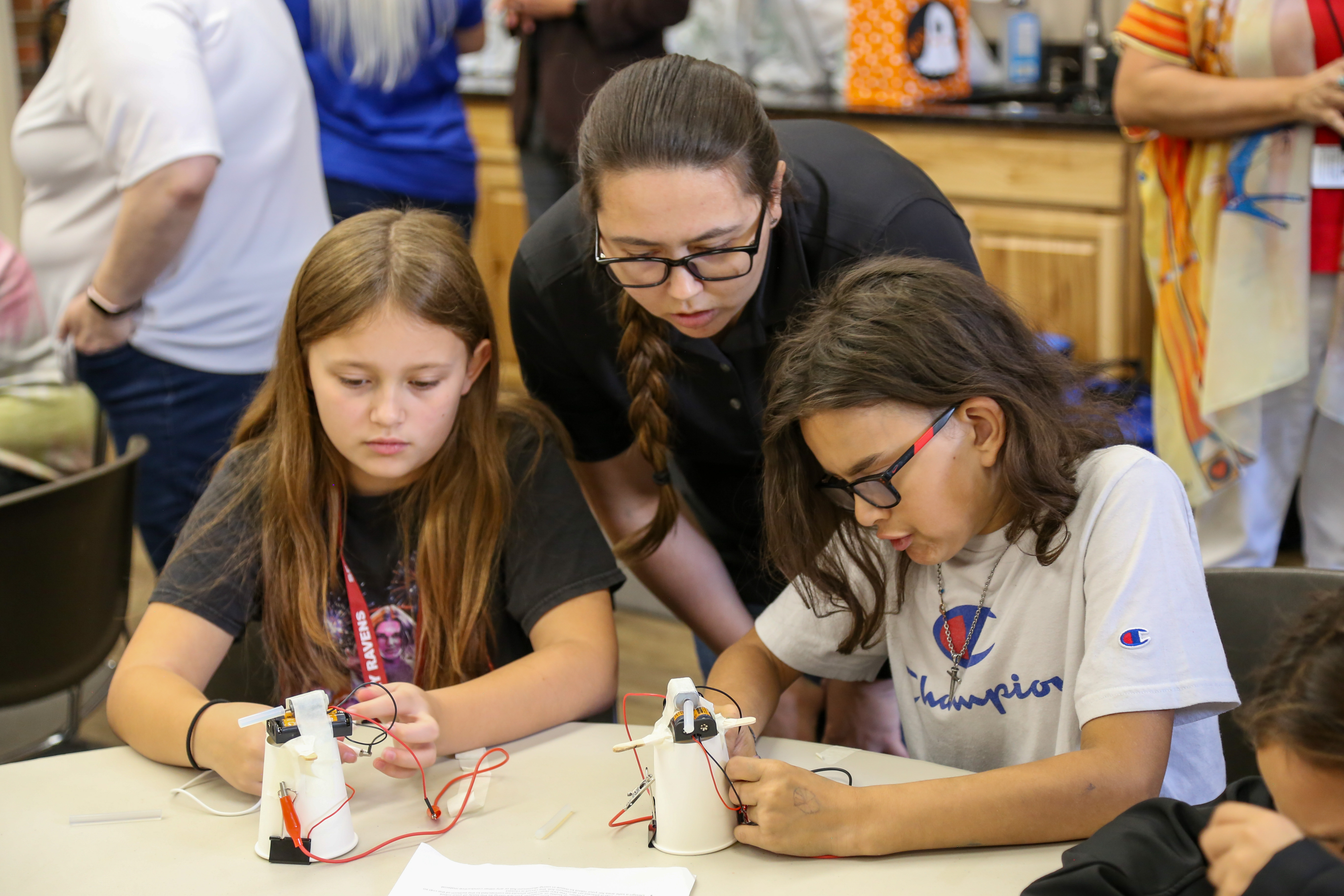Casey Denham, a Meskwaki woman, leans over to help two tribal students with a STEM project. The children sit at a white round table. Each of them holds wires in their hands. The black and red wires are connected to paper cups.