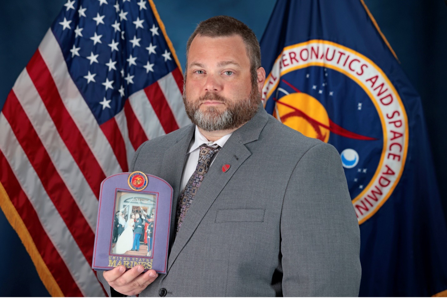 A man in a grey suit holds a photo of his wedding day wearing his military uniform in front of an American flag and a NASA flag.