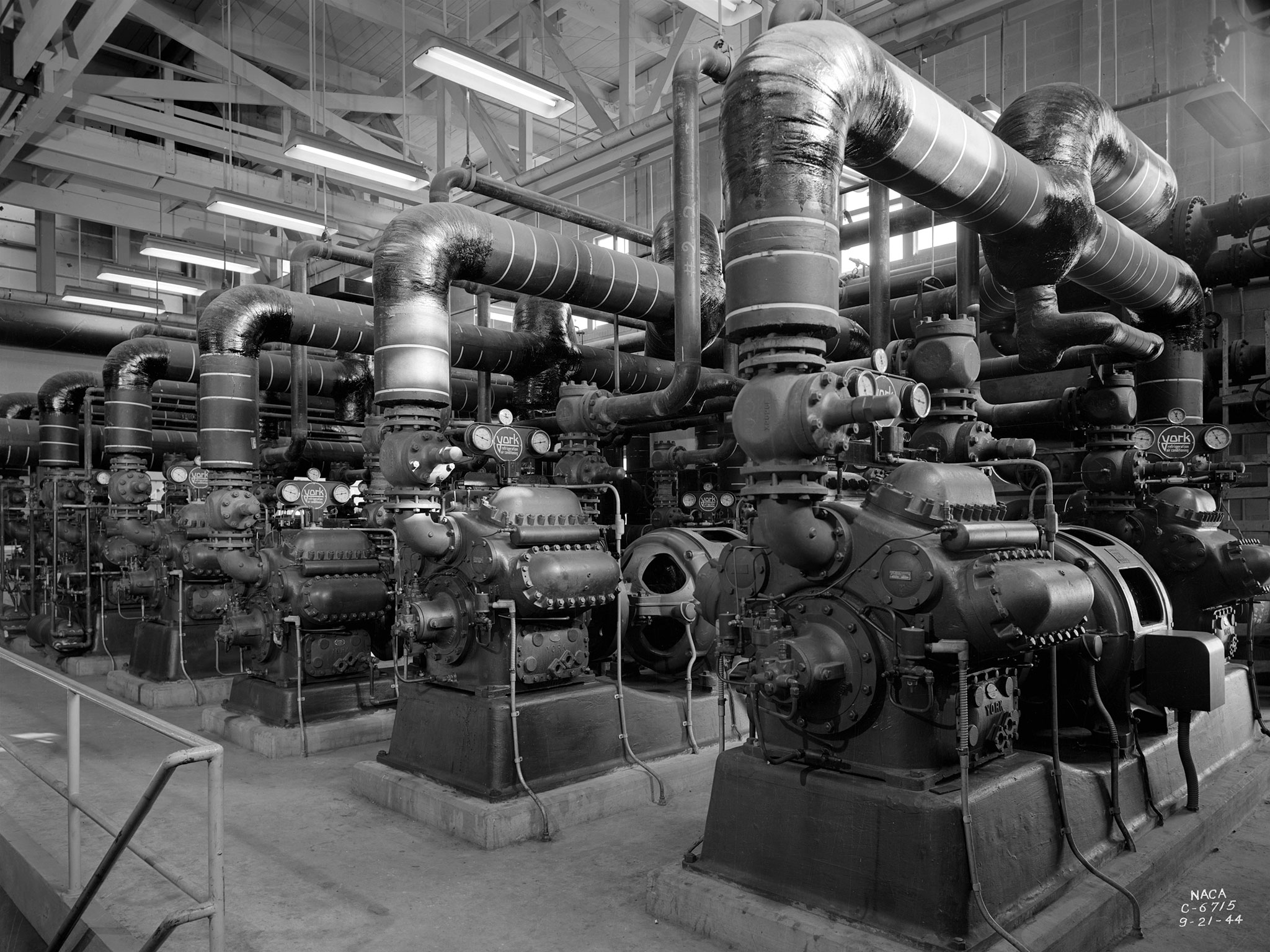 A black-and-white photo of a row of large compressors inside a warehouse building. They are metal and industrial with many gauges, tubes, and valves attached.