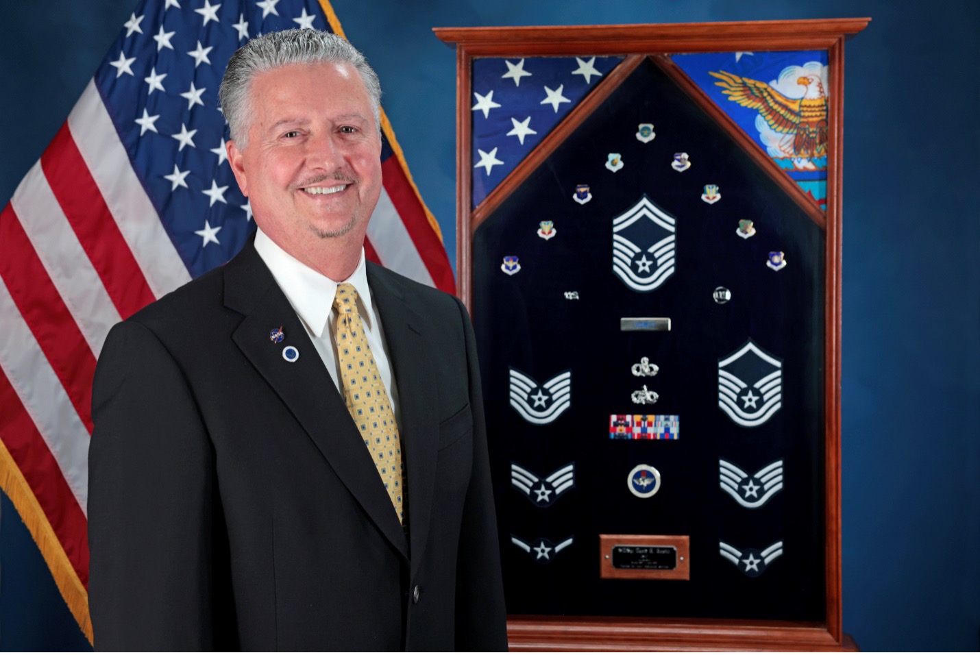 A man wearing a black suit stands with his shadow box full of military medals in front of an American Flag.
