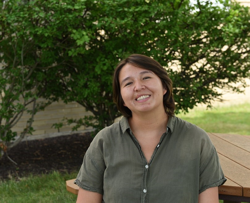 Alyssa Warrior poses outdoors and smiles at the camera. She is sitting at a wooden picnic table in front of a bright green bush. She is wearing a dark green collared shirt.