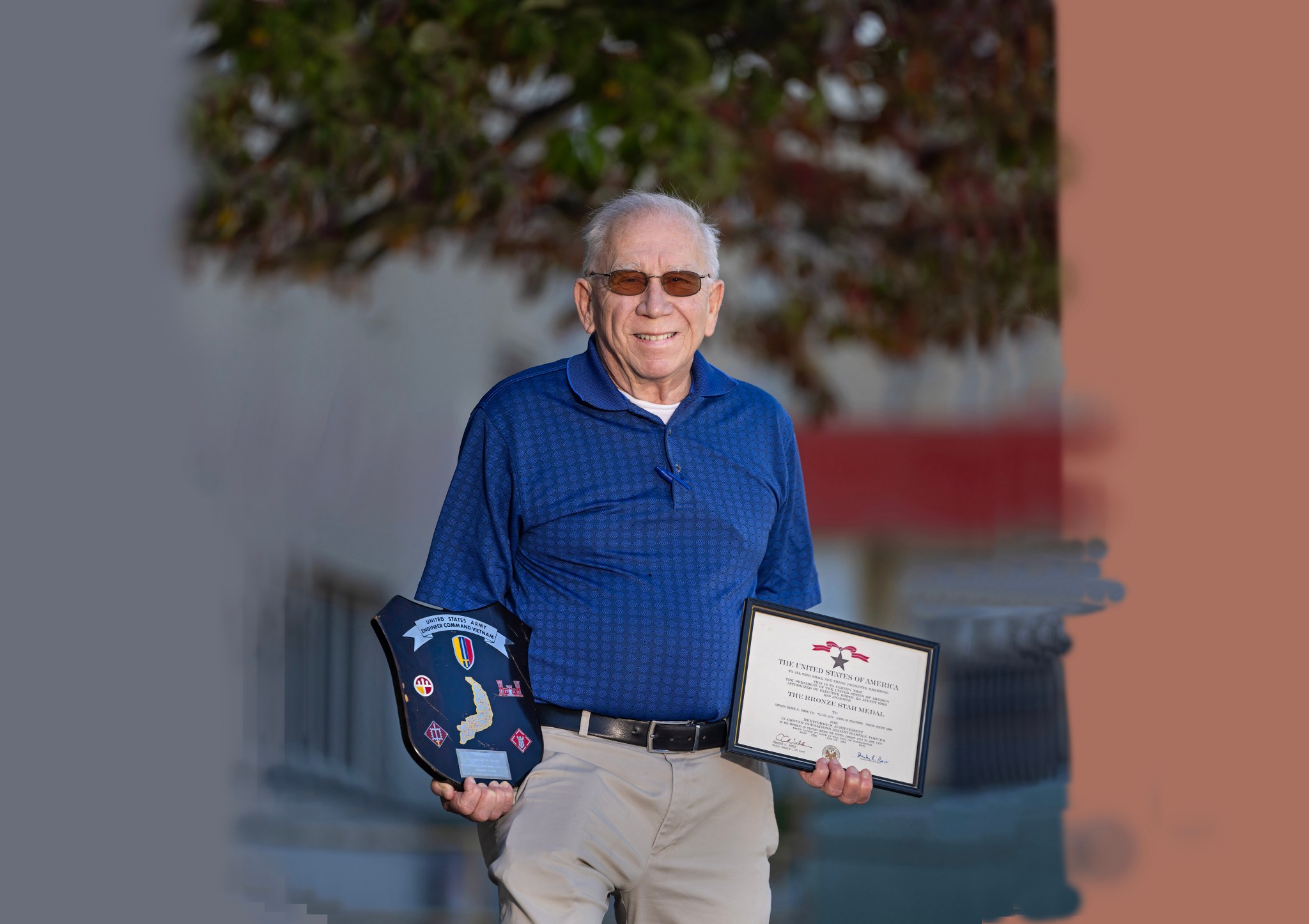 An older white man stands in front of a tree holding two plaques.