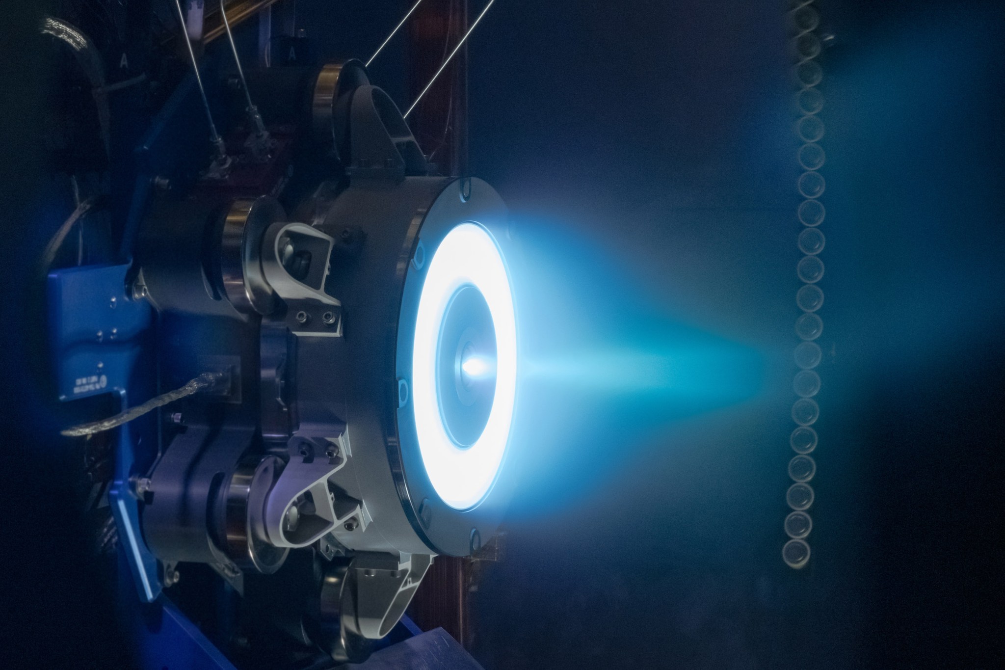 A solar electric propulsion thruster emits the blue hue of Xenon gas during testing. Vibrant blue light emanates in a circular shape from the dark grey thruster, which is mounted inside a vacuum chamber. The blue light then narrows into a plume as it moves farther away from the thruster, illuminating the otherwise darkened chamber.