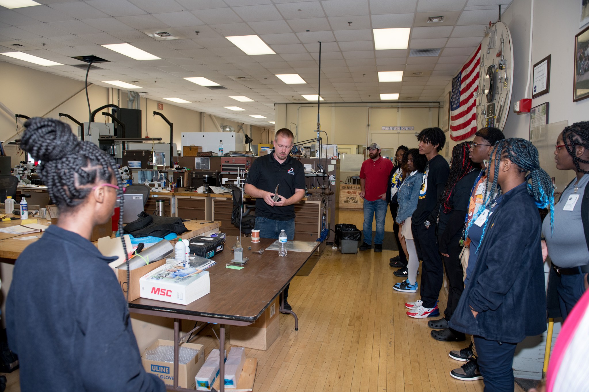 Students stand and watch as Chris Metro who is holding a steel part designed in NASA Glenn Research Center’s Manufacturing Facility.