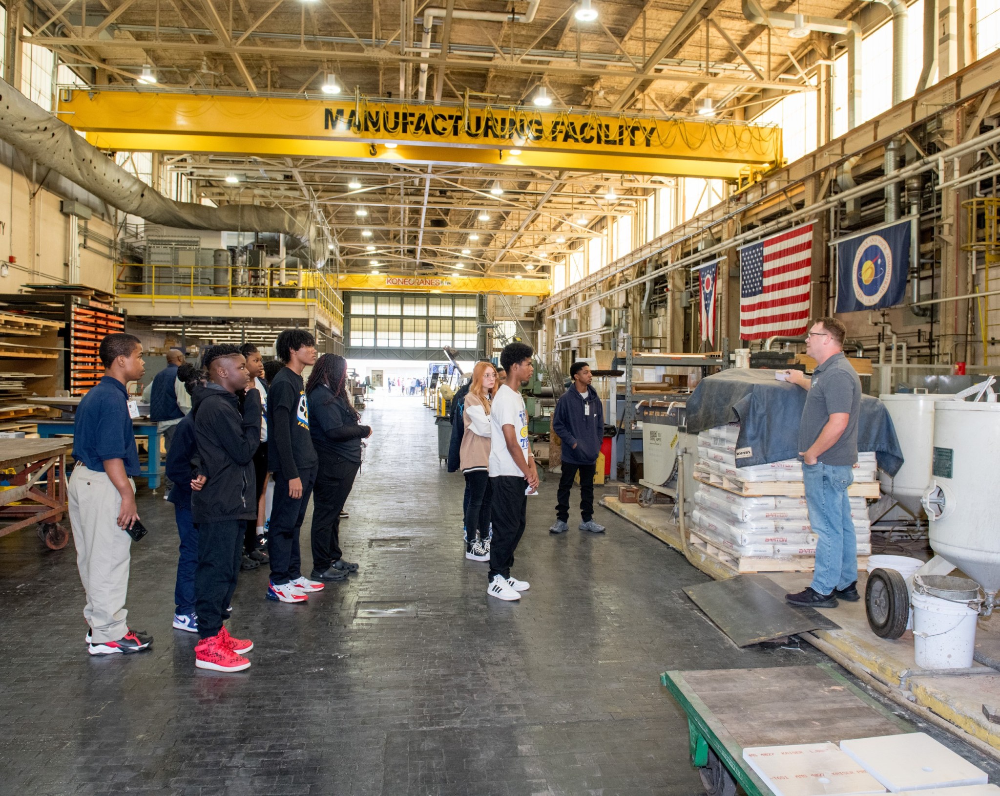 Inside a large warehouse-type facility, students observe Matt Conley as he talks about manufacturing at NASA’s Glenn Research Center. 
