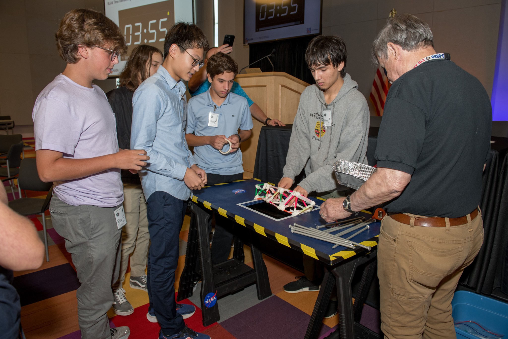 lt Text: Students gather around a small table to build a truss made from straws.