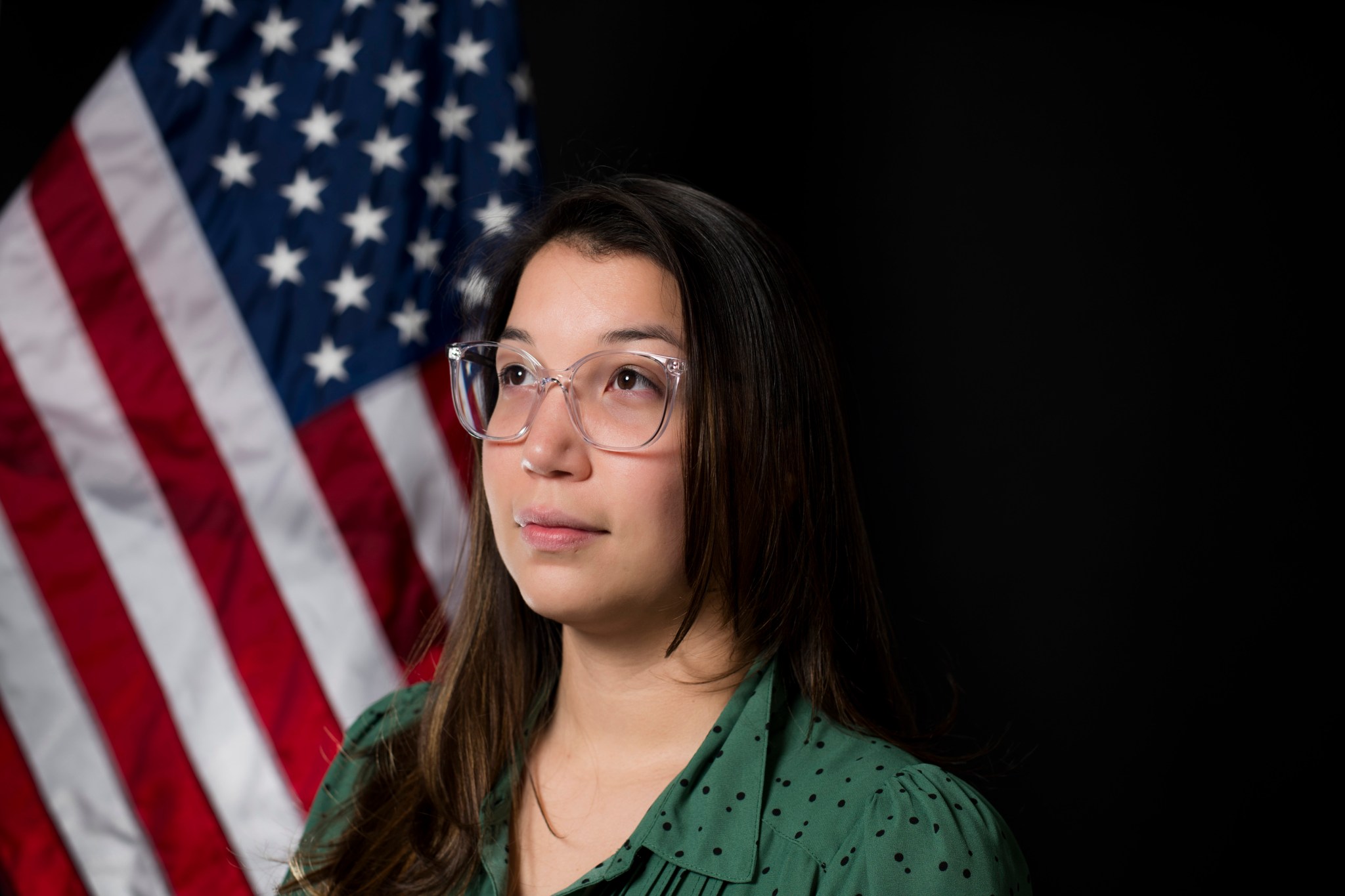 Sydney Khamphoune poses in front of an American flag. She is wearing a green shirt with clear glasses and is looking into the distance to the left of the camera.