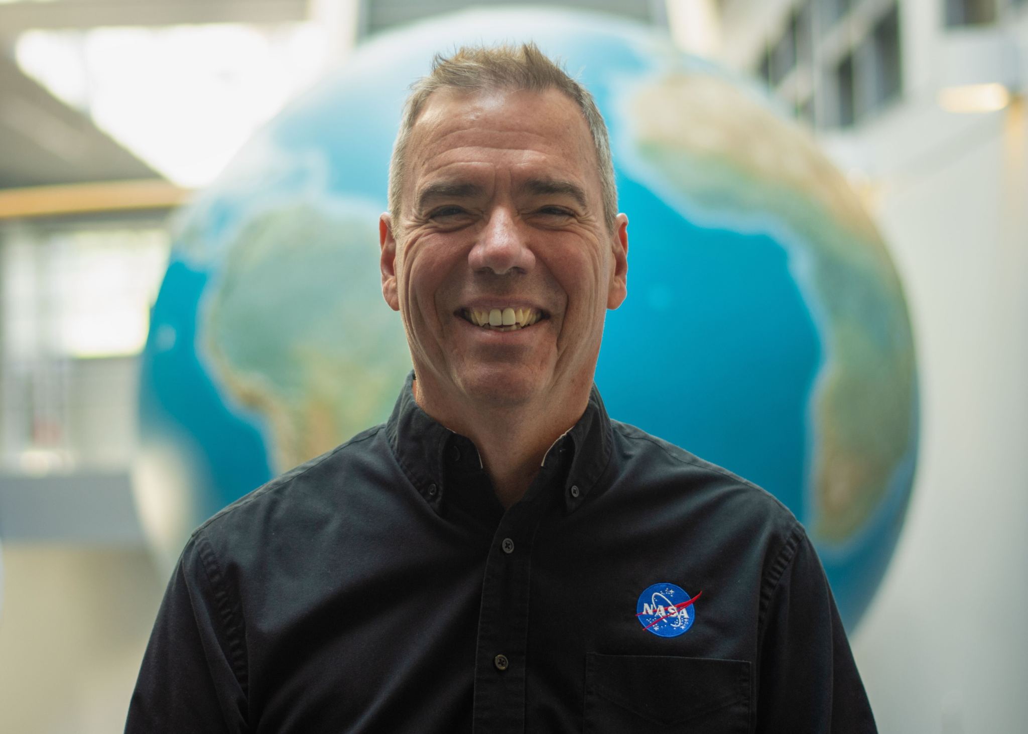 Peter Griffith, a man with short gray hair, smiles and poses for an official portrait. He wears a black collared shirt with the NASA logo on his left chest. A huge model of Earth is visible out of focus behind him in a large, sunlit room.