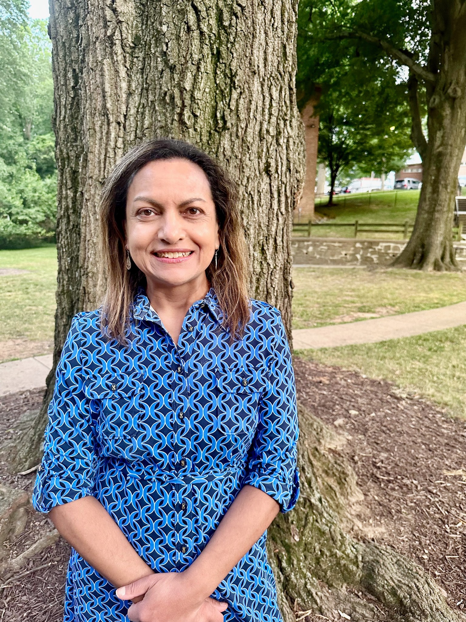Rita Owens, a woman with shoulder-length, light brown hair, smiles at the camera in a casual outdoor portrait. She wears a patterned blue dress and stands in front of a tree. More green trees, a stone wall and brick buildings with cars are visible in the background.