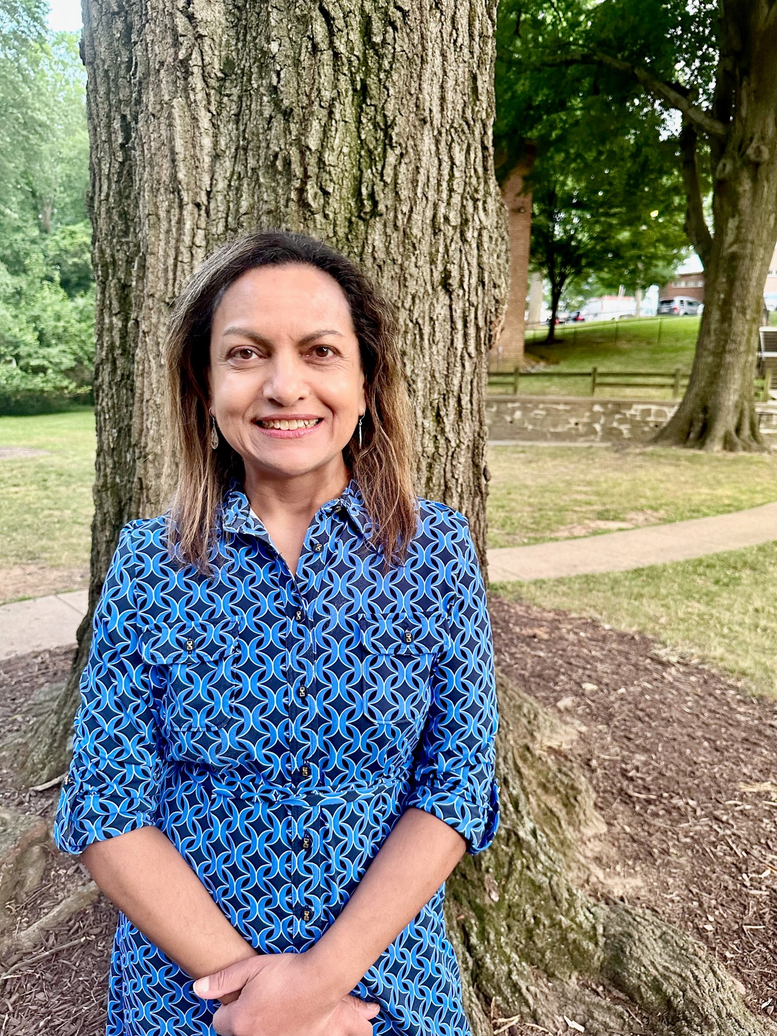 Rita Owens, a woman with shoulder-length, light brown hair, smiles at the camera in a casual outdoor portrait. She wears a patterned blue dress and stands in front of a tree. More green trees, a stone wall and brick buildings with cars are visible in the background.