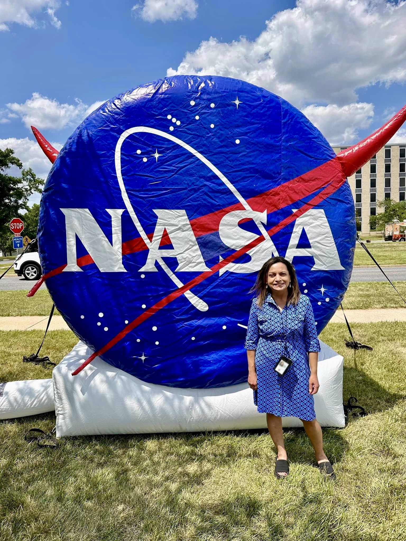 Rita Owens, a woman with shoulder-length, light brown hair, smiles and stands in front of a large inflatable shaped like the NASA logo. It is a large, bright blue circle dotted with stars and crossed by a red, V-shaped swoosh. 