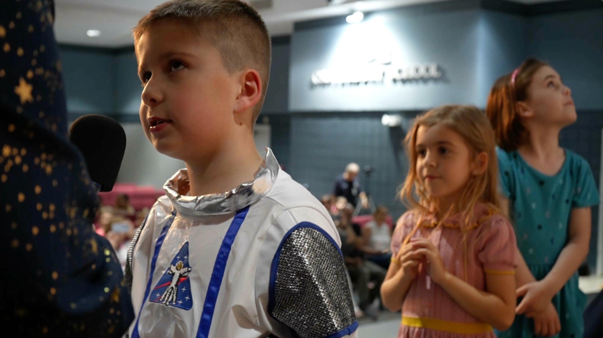 Student Isaac Deeter, wearing an astronaut costume, speaks into a microphone. Samantha Pezzi, wearing a pink dress, her hands in front of her, waits behind him and a student in a blue dress stands behind her. The school auditorium is blurred in the background.