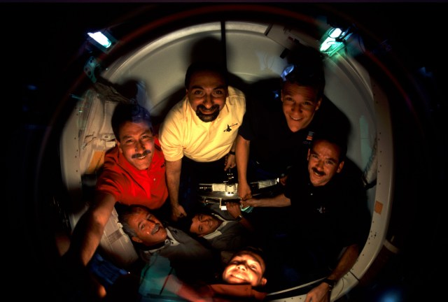 The six astronauts and one cosmonaut comprising the STS-100 crew assemble in the Pressurized Mating Adapter (PMA-2) while waiting to visit the Expedition Two crew and the International Space Station (ISS). With his arm extended to left foreground is astronaut Kent V. Rominger, STS-100 mission commander. In the circular arrangement of crew members, clockwise from Rominger's position, are astronauts Umberto Guidoni, Scott E. Parazynski, Chris A. Hadfield, Jeffrey S. Ashby and John L. Phillips. Cosmonaut Yuri V. Lonchakov's head emerges at bottom center. On the other side of the glass were the Expedition Two crew members--cosmonaut Yury V. Usachev and astronauts James S. Voss and Susan J. Helms. Lonchakov and Usachev represent Rosaviakosmos; Hadfield is with the Canadian Space Agency (CSA) and Guidoni is associated with the European Space Agency (ESA). The ten were beginning a day that went on to see the first opening of hatches linking the two spacecraft, an impressive first step by the station’s new Canadarm2 and the berthing to the station of Raffaello, the Italian-built logistics module. Hatch opening was set for 4 a.m (CDT), April 23.