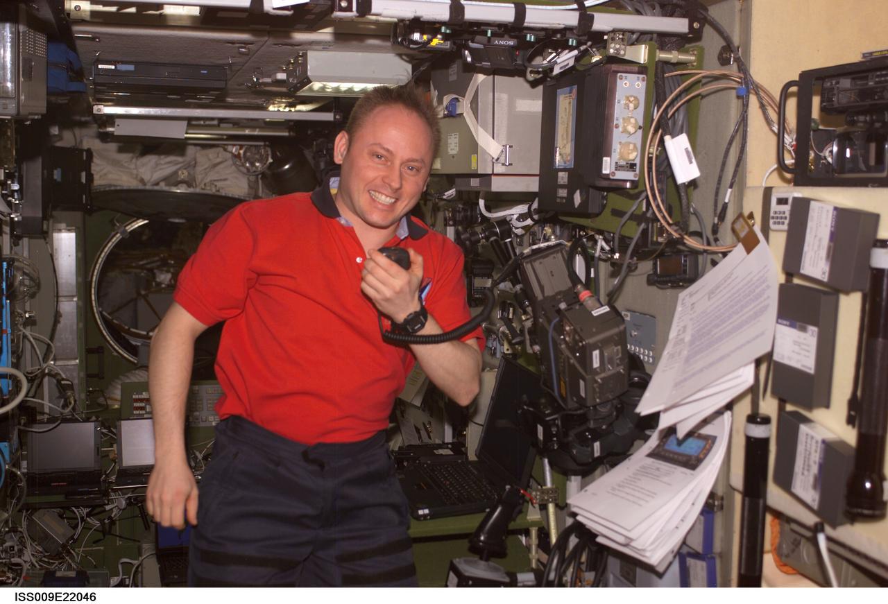 Mike Fincke wears a red shirt and smiles at the camera as he holds the ham radio mic in his left hand next to the radio set. Sheafs of paper are clipped to the wall in front of him and equipment covers the wall behind him.